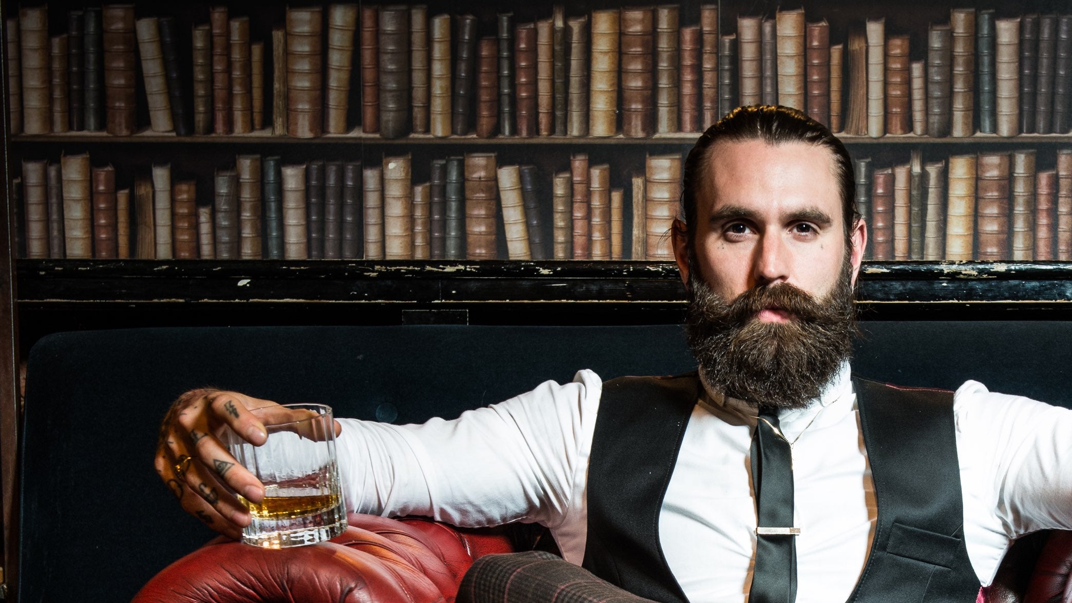 Mann i 30-årene med skjegg som sitter i en sofa foran en bokhylle og har en drink i hånden